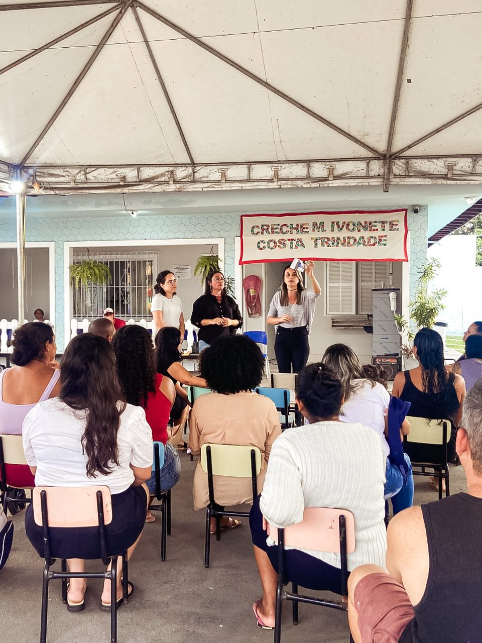 Três integrantes da Rebrapi conversando com famílias sentadas em cadeiras, sob uma tenda, em frente à Creche M. Ivonete Costa Trindade