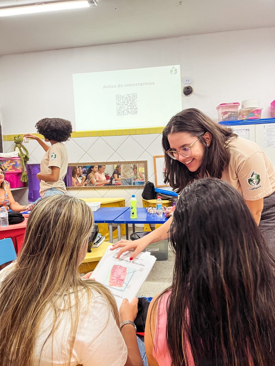 Integrante da Rebrapi mostrando um material educativo a duas estudantes durante uma ação educativa