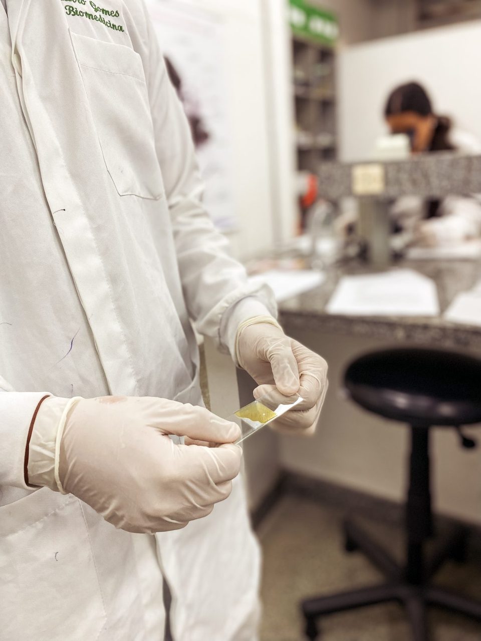 Pessoa de jaleco e luvas segurando uma lâmina no laboratório